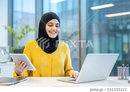 Smiling businesswoman wearing a hijab, working at a desk in a modern office, using a laptop and digital tablet to manage tasks and communicate effectively in a professional environment Smiling businesswoman wearing a hijab, working at a desk in a modern office, using a laptop and digital tablet to manage tasks and communicate effectively in a professional environment 132335122