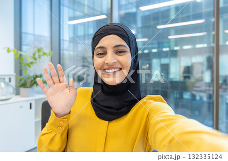 Muslim businesswoman wearing a hijab, smiling and waving hand to the camera during a video conference or looking at a selfie in a modern corporate office setting 132335124