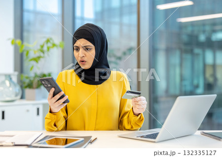 Muslim businesswoman wearing a hijab expressing shock and frustration while looking at her smartphone and holding a credit card, experiencing an online shopping payment error or fraud 132335127