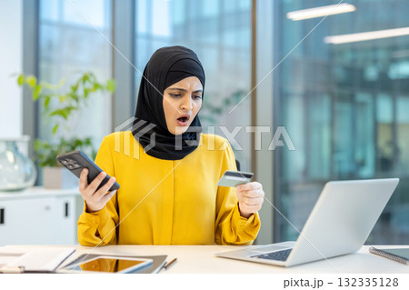 Arabian woman wearing a hijab sitting at an office desk, holding a credit card and smartphone, showing a shocked expression due to an online shopping payment problem or fraud 132335128