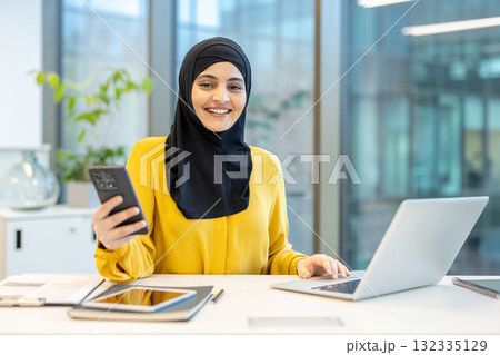 Young muslim businesswoman wearing a hijab is working at her desk in a modern office, holding a smartphone and using a laptop while smiling Young muslim businesswoman wearing a hijab is working at her desk in a modern office, holding a smartphone and using a laptop while smiling 132335129