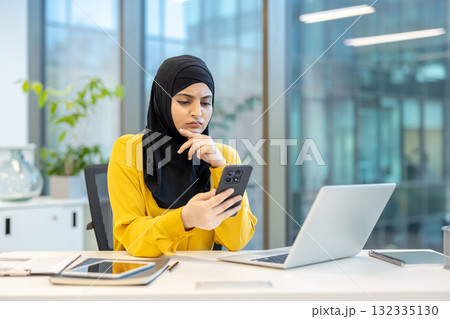 Muslim businesswoman wearing a hijab sitting at her modern office desk, reading her smartphone with a concerned and thoughtful expression while working on her laptop Muslim businesswoman wearing a hijab sitting at her modern office desk, reading her smartphone with a concerned and thoughtful expression while working on her laptop 132335130
