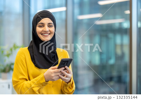 Muslim businesswoman smiling towards camera while holding and using a smartphone, standing in a modern corporate office environment, representing technology, communication, and professional success 132335144