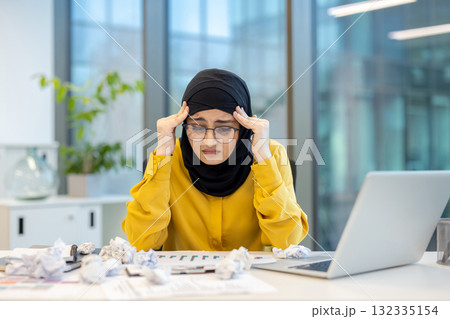 Young muslim businesswoman in hijab and glasses clutching her temples at a cluttered office desk, stressed and fatigued from overwork, missed deadlines and burnout Young muslim businesswoman in hijab and glasses clutching her temples at a cluttered office desk, stressed and fatigued from overwork, missed deadlines and burnout 132335154