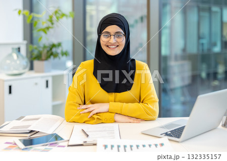 Young muslim businesswoman in hijab and eyeglasses smiling positively while sitting at a desk in a contemporary office setting, representing professionalism and success 132335157