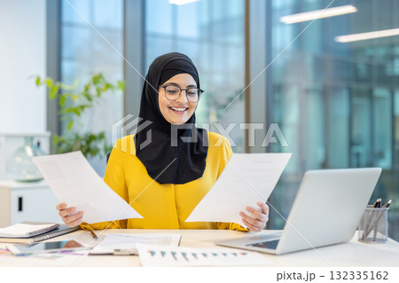 Muslim businesswoman wearing hijab and eyeglasses, happily reviewing business documents while working at her desk in a contemporary office environment 132335162