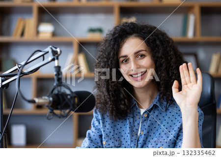 Young woman vlogger with curly hair smiling and waving to camera while presenting a podcast and streaming from a cozy modern home studio ,with microphone and bookshelf Young woman vlogger with curly hair smiling and waving to camera while presenting a podcast and streaming from a cozy modern home studio ,with microphone and bookshelf 132335462