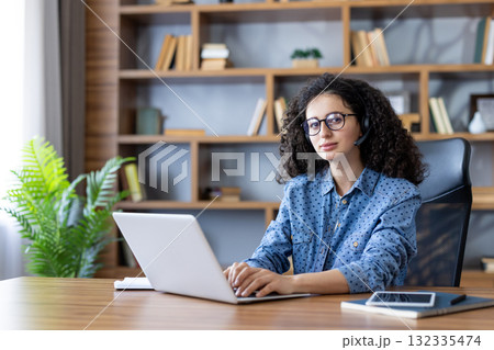 Young woman with curly hair and glasses wearing a headset, focused on laptop at a home office desk providing customer support and virtual assistance during remote work Young woman with curly hair and glasses wearing a headset, focused on laptop at a home office desk providing customer support and virtual assistance during remote work 132335474