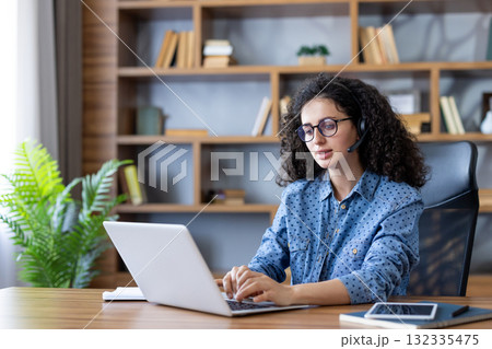 Young woman with curly hair wearing a headset with a microphone. Concentrating while typing on a laptop. Working remotely from her modern home office setup with a bookshelf and plant in the background 132335475