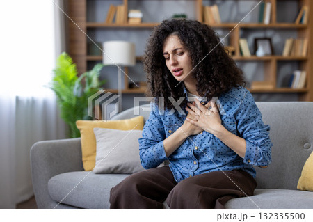 Young distressed woman feeling sudden sharp chest pain and difficulty breathing, suffering from a panic attack or heart problems while sitting on a couch at home Young distressed woman feeling sudden sharp chest pain and difficulty breathing, suffering from a panic attack or heart problems while sitting on a couch at home 132335500