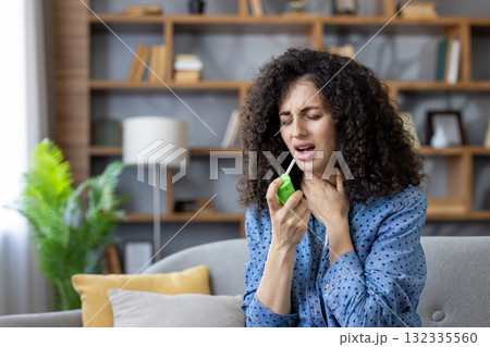 Young woman on a couch spraying throat medicine into her mouth, wincing in discomfort as she self-treats sore throat and cold symptoms at home 132335560