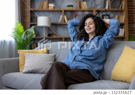 Young woman enjoying a moment of peace and relaxation on her comfortable sofa, hands behind her head, eyes closed, a peaceful smile on her face, surrounded by the cozy atmosphere of her living room 132335627