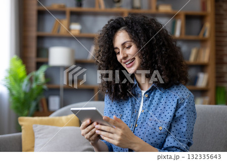 Woman with curly hair smiling and looking at a tablet, sitting on a sofa at home, enjoying online content and connectivity for entertainment and relaxation Woman with curly hair smiling and looking at a tablet, sitting on a sofa at home, enjoying online content and connectivity for entertainment and relaxation 132335643