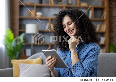 Happy young woman with curly hair relaxing on a sofa, smiling while browsing content on a digital tablet, enjoying leisure time in a modern living room with a bookshelf 132335644