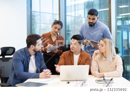 A business woman in a yellow blouse and hijab sits at her desk in an office, holding documents and a pen. A business woman in a yellow blouse and hijab sits at her desk in an office, holding documents and a pen. 132335692