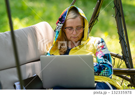 senior woman with laptop and documents working in garden on rocking couch, green home office concept 132335978