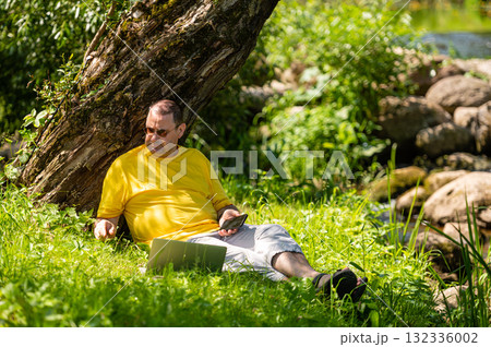 man with laptop and smartphone working outside in the meadow by the river, outdoor office concept man with laptop and smartphone working outside in the meadow by the river, outdoor office concept 132336002