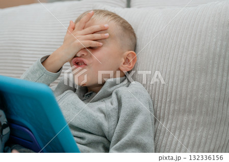 Young boy sitting on a couch with a tablet, covering his face with his hand in a playful facepalm candid moment of frustration Young boy sitting on a couch with a tablet, covering his face with his hand in a playful facepalm candid moment of frustration 132336156