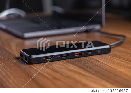 Black USB-C hub with multiple HDMI, USB, and PD ports connected to a laptop on a wooden desk, optimizing modern digital workspace connectivity for productivity 132336427