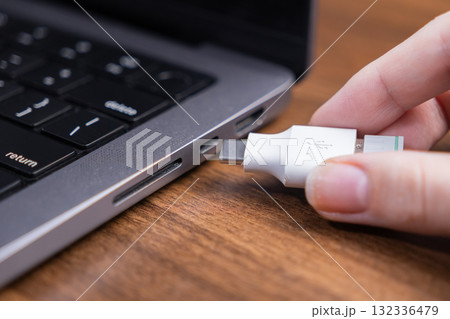 A hand inserts a white USB Type-C adapter into a silver laptop port on a wooden desk, emphasizing modern device connectivity for data transfer and charging 132336479