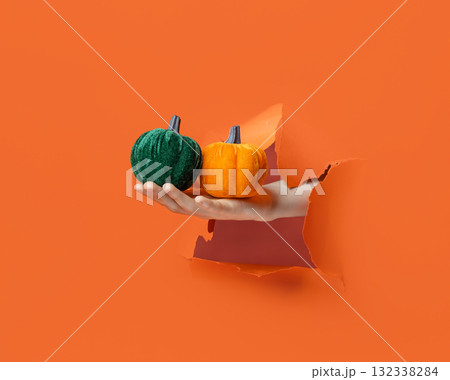 Hand holding soft wool pumpkin with golden stem through torn orange paper background. 132338284