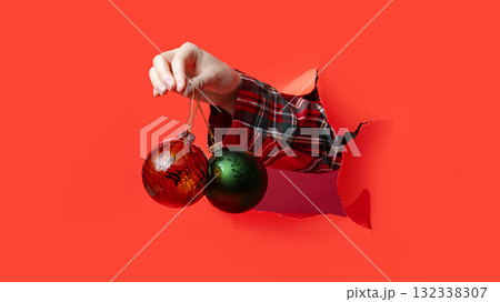 Merry Christmas, hand holds a set of three shiny Christmas tree baubles green, red, and pink through torn red background. Merry Christmas, hand holds a set of three shiny Christmas tree baubles green, red, and pink through torn red background. 132338307