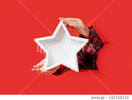 Hands in plaid shirt hold white star-shaped tray emerging through torn red paper, against festive red background. Hands in plaid shirt hold white star-shaped tray emerging through torn red paper, against festive red background. 132338310