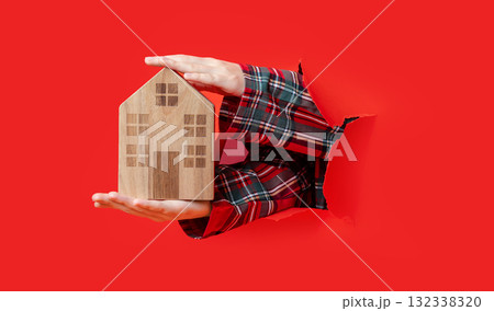 Merry Christmas. hand holding wooden toy house and pointing at it through torn red paper, plaid shirt visible. Merry Christmas. hand holding wooden toy house and pointing at it through torn red paper, plaid shirt visible. 132338320