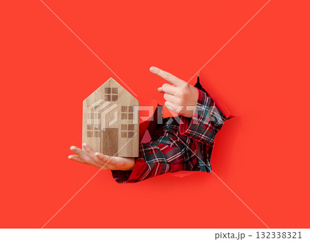 Merry Christmas. hand holding wooden toy house and pointing at it through torn red paper Merry Christmas. hand holding wooden toy house and pointing at it through torn red paper 132338321