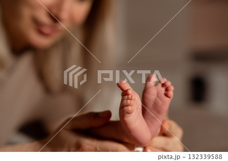Mother gently holding her newborn babys tiny feet while smiling in a cozy indoor setting during early morning light 132339588