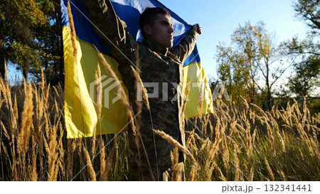 Military man in uniform stands with raised over head flag of Ukraine at countryside. Male ukrainian army soldier with lifted national banner in honor of victory against russian aggression. End of war 132341441