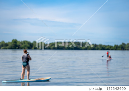 People paddle boarding on a calm lake under a clear blue sky during the summer afternoon in a peaceful outdoor setting 132342490