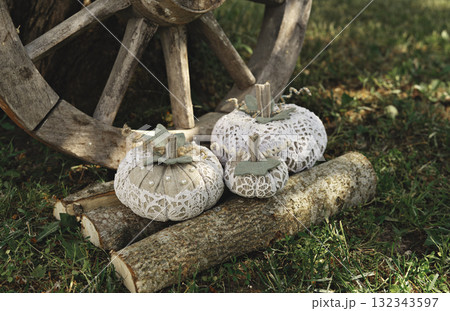 Handmade lace fabric pumpkins rest on cut logs near old wooden wagon wheel, rustic outdoor garden scene with cozy autumn mood 132343597