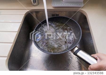 Hand rinses oily frying pan under faucet in stainless sink, unsafe contact of water with hot oil and visible splashes Hand rinses oily frying pan under faucet in stainless sink, unsafe contact of water with hot oil and visible splashes 132343598