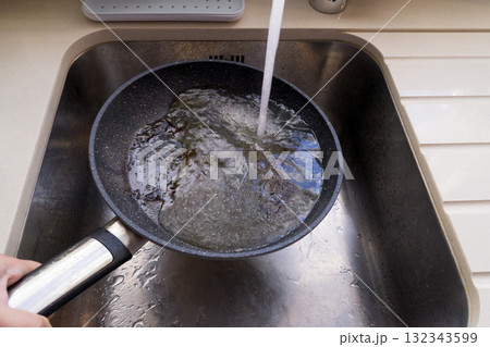 Hand holds frying pan with hot oil under faucet in stainless sink, water hitting oil creates risky splash and steam 132343599