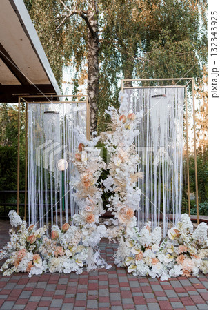 Romantic Wedding Ceremony Backdrop with White Floral Arrangement, String Curtains, Golden Accents, and Outdoor Garden Setting at Dusk. 132343925