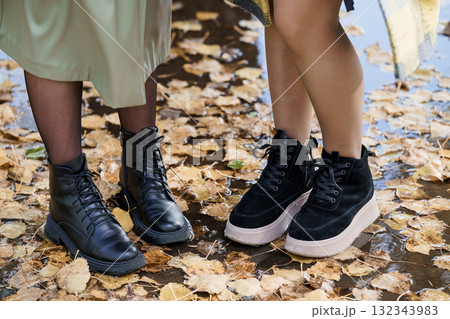Autumn Fashion Boots Two Womens Legs and Black Leather Ankle Boots Standing in Fallen Leaves, Outdoor Style, Fall Colors, Close-Up Footwear. Autumn Fashion Boots Two Womens Legs and Black Leather Ankle Boots Standing in Fallen Leaves, Outdoor Style, Fall Colors, Close-Up Footwear. 132343983