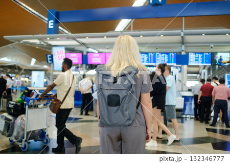 Busy airport check-in area with travelers waiting at counters, focusing on a woman with a backpack and others in the background during the day 132346777
