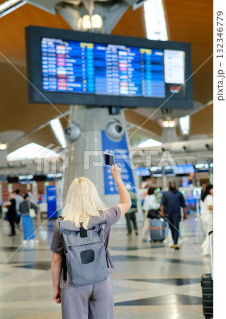 Passenger checks flight details at an airport terminal while holding a passport in the air and preparing for travel during a sunny day 132346779
