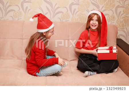 Girls in New Year's looks with a gift are sitting at home on the sofa. 132349923
