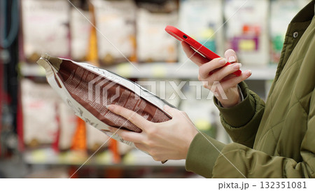 Woman scanning dry pet food with smartphone in supermarket. Modern customer checking product information and ingredients using mobile app, AI assistant. Smart shopping Digital retail Conscious choice 132351081