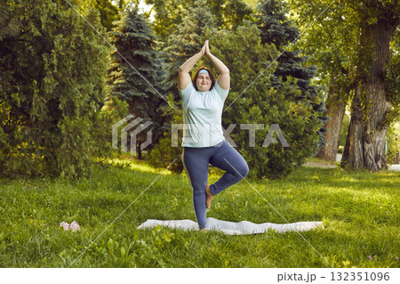 Happy overweight woman is doing yoga in nature, standing in Tree Pose and relaxing 132351096