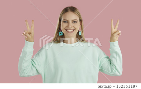Blondy happy girl in mint sweatshier showing victory signs by two hands on pink background. Blondy happy girl in mint sweatshier showing victory signs by two hands on pink background. 132351197
