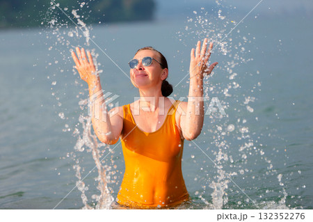 An adult woman splashes sea water on herself. She is wearing a yellow wet T-shirt and sunglasses. 132352276