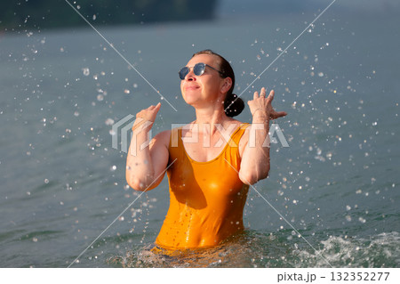 An adult woman splashes sea water on herself. She is wearing a yellow wet T-shirt and sunglasses. 132352277