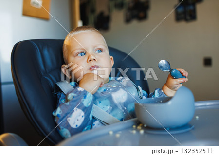 Cute one-year-old boy sits in a high chair and eats independently. Silicone utensils for feeding children. 132352511
