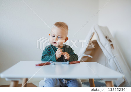 Toddler boy in a bright room sits at a children's table and plays with toys. Furniture for children. Table and chairs for babies. Child development 132352564