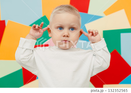 Happy little toddler in a white t-shirt and jeans stands on a studio background and covers his ears, expressing joy and curiosity. Stylish toddler. Concept of parenthood, natural beauty, love and care 132352565