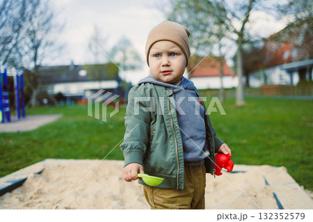 A boy of 2 years old in a spring jacket and hat plays with sand in a sandbox. A boy plays on a playground with a shovel in the sand. A child's walk in the fresh air 132352579