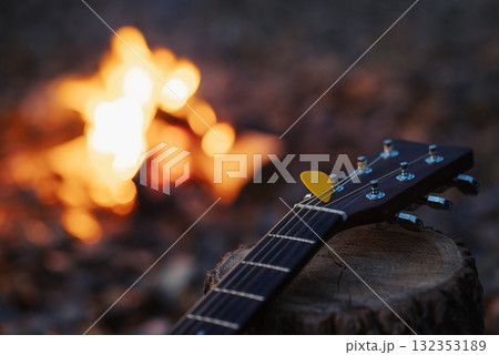 Close up of guitar on stump with bonfire on the background. Neck of guitar, yellow mediator on wooden stump near campfire Close up of guitar on stump with bonfire on the background. Neck of guitar, yellow mediator on wooden stump near campfire 132353189
